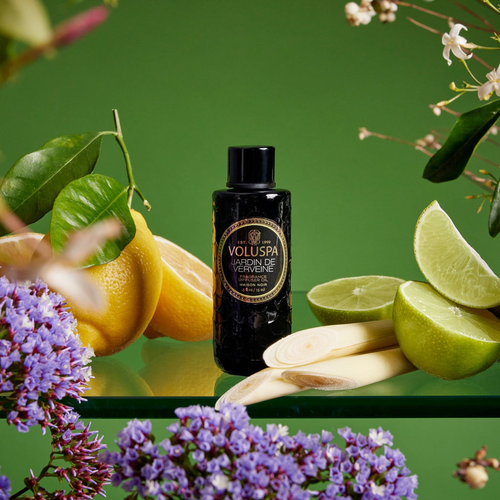 A Voluspa Jardin De Verveine Blanc Ultrasonic Diffuser Fragrance Oil bottle stands amidst fresh lemons, limes, and lemongrass on glass, with purple and white flowers framing the scene against a lush green backdrop.