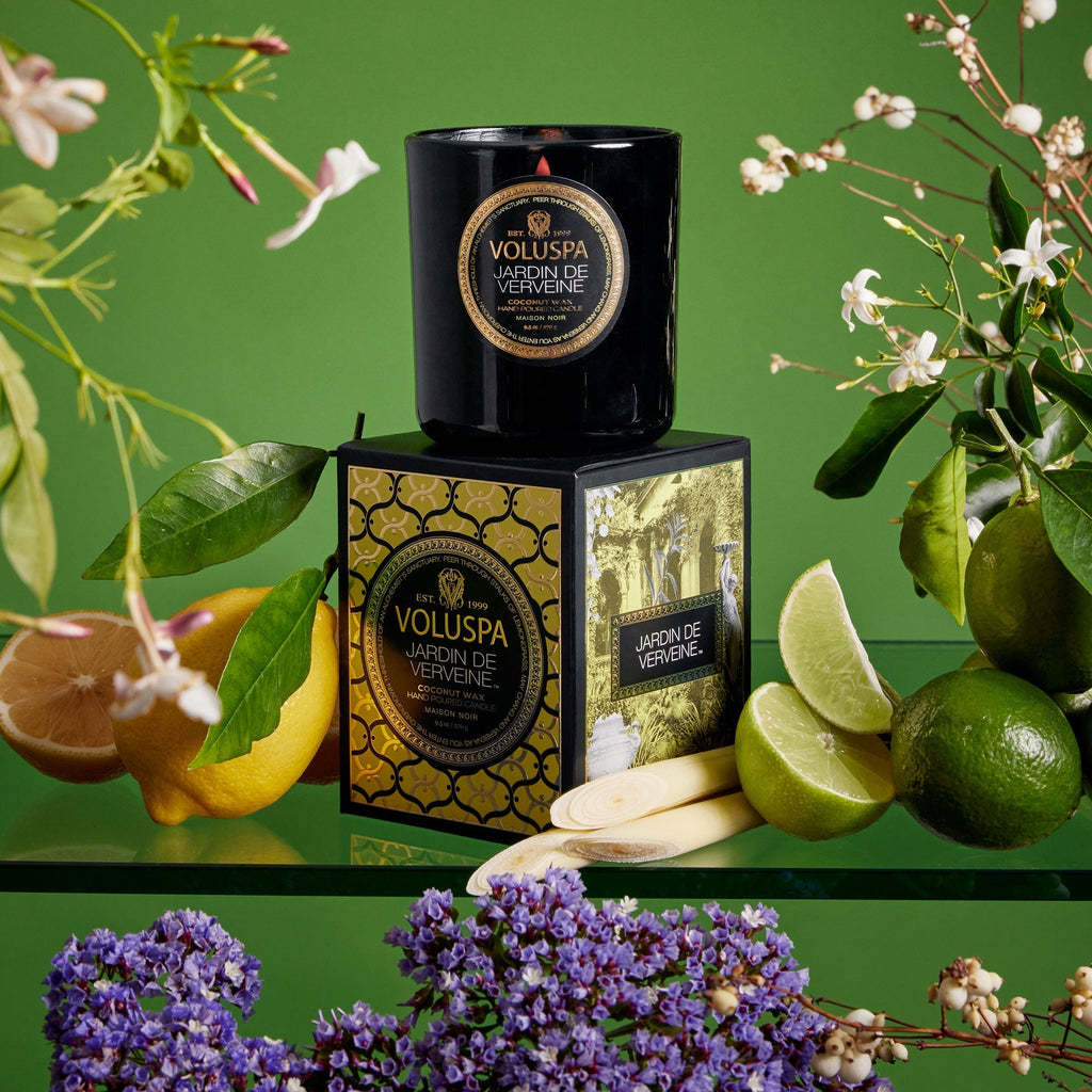 A Voluspa Jardin De Verveine Classic Candle 9.5oz, made with a coconut wax blend, is displayed on a decorative box amidst lemongrass, lime halves, lemons, and white flowers. The green backdrop highlights the fresh setting and enhances its fragrance throw.