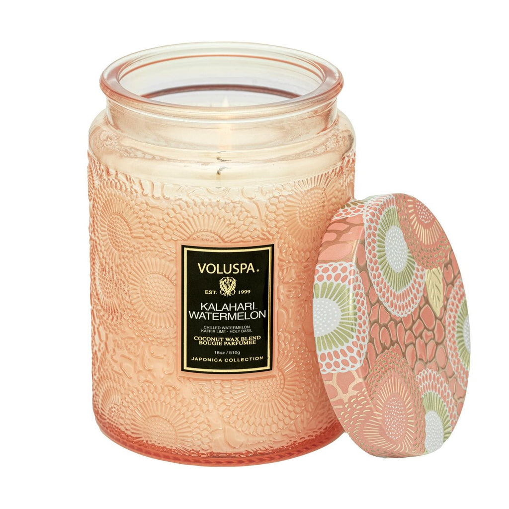 A textured glass jar candle labeled Voluspa Kalahari Watermelon rests beside its floral-patterned lid in pink, green, and white. Featuring natural wicks and a peach-toned coconut wax blend, it exudes chilled watermelon notes.