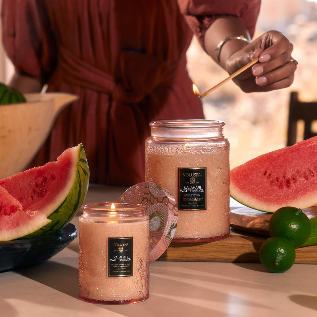 Someone lights the Voluspa - Kalahari Watermelon Large Jar 18oz Candle on a table. Made from a coconut wax blend, its glow complements chilled watermelon slices and whole limes, enhancing the serene summer scene with a softly blurred background for added tranquility.
.