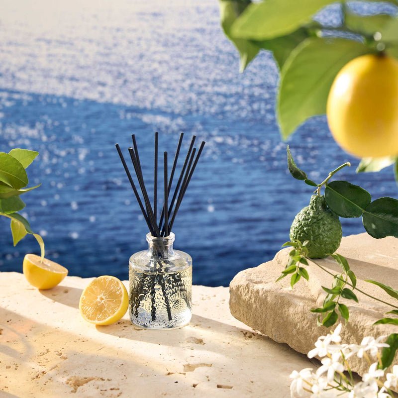 The Voluspa Mediterranean Lemon 3.4oz Reed Diffuser, featuring black reeds, rests on a sunlit stone with sliced lemons and white flowers, filling the air with a fresh citrus scent reminiscent of the sparkling Mediterranean Sea.