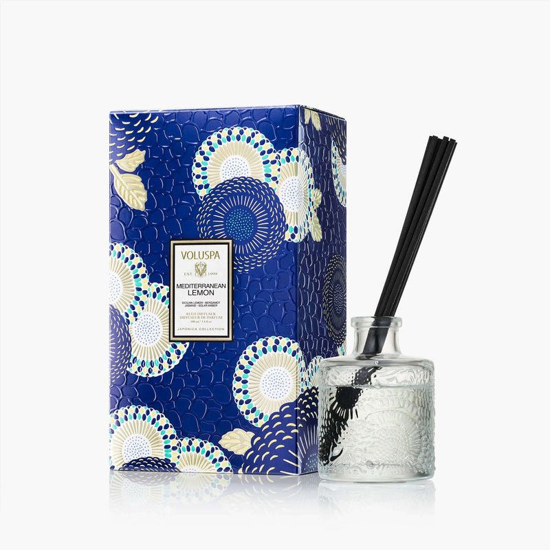 The Voluspa Mediterranean Lemon 3.4oz Reed Diffuser features a clear textured glass bottle with black reeds and comes in a blue and white patterned box, filling your space with a refreshing citrus scent.