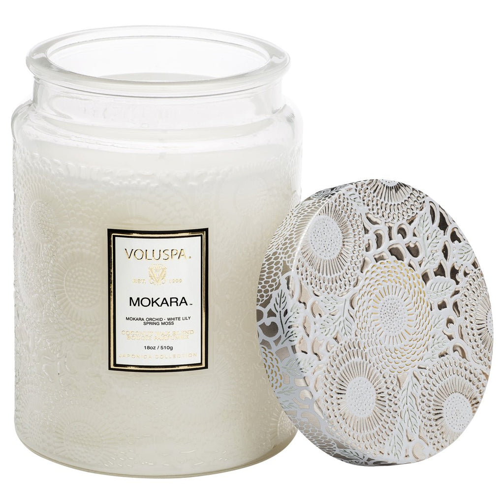 An 18oz Voluspa Mokara Large Jar Candle, elegantly textured with a floral-patterned white lid, features coconut wax that highlights its Mokara orchid, white lily, and spring moss fragrance.