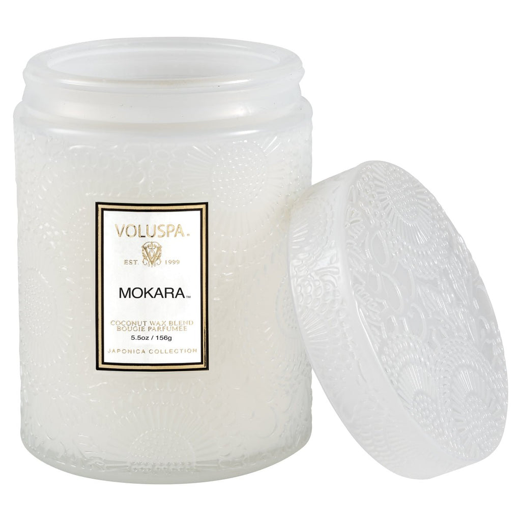 The Voluspa - Mokara Small Jar 5.5oz Candle features a frosted glass design with a removable white lid, showcasing floral patterns reminiscent of an island garden. Filled with coconut wax, it embodies the delicate Mokara Orchid essence as detailed on its label.