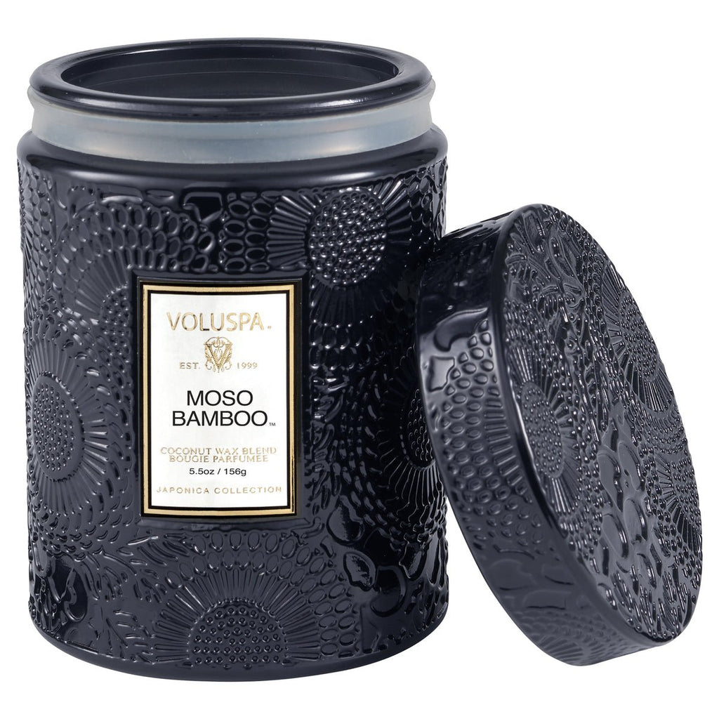 A Voluspa Moso Bamboo small jar candle (5.5 oz) features a black flower-patterned design with its lid aside. Part of the Japonica Collection, it contains a coconut wax blend with Japanese Cypress notes.