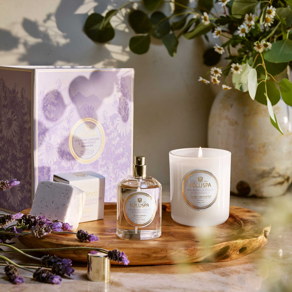 A wooden tray holds the Voluspa - Mountain Lavender & Chamomile Retreat Set with a white candle, fragrance spray, bath bomb, and Provence Lavender box. Accents include lavender sprigs, greenery, and a ceramic vase with small white flowers.