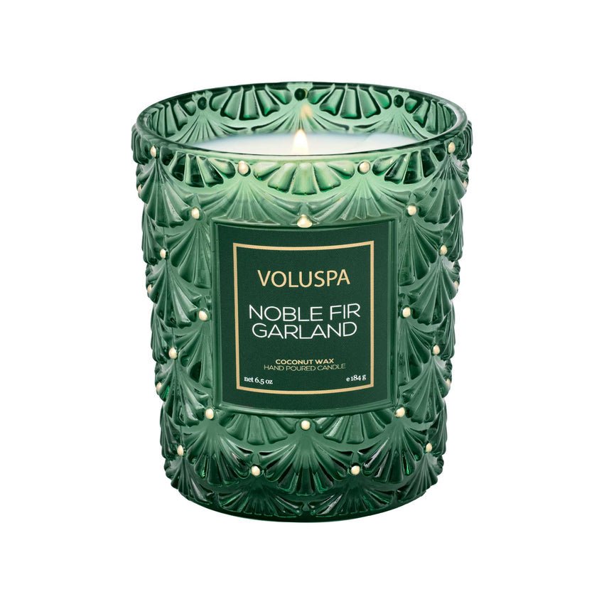 The Voluspa Noble Fir Garland Classic 6.5oz Candle features a woody scent and comes in a decorative glass jar with an embossed leaf pattern and pearl-like accents.