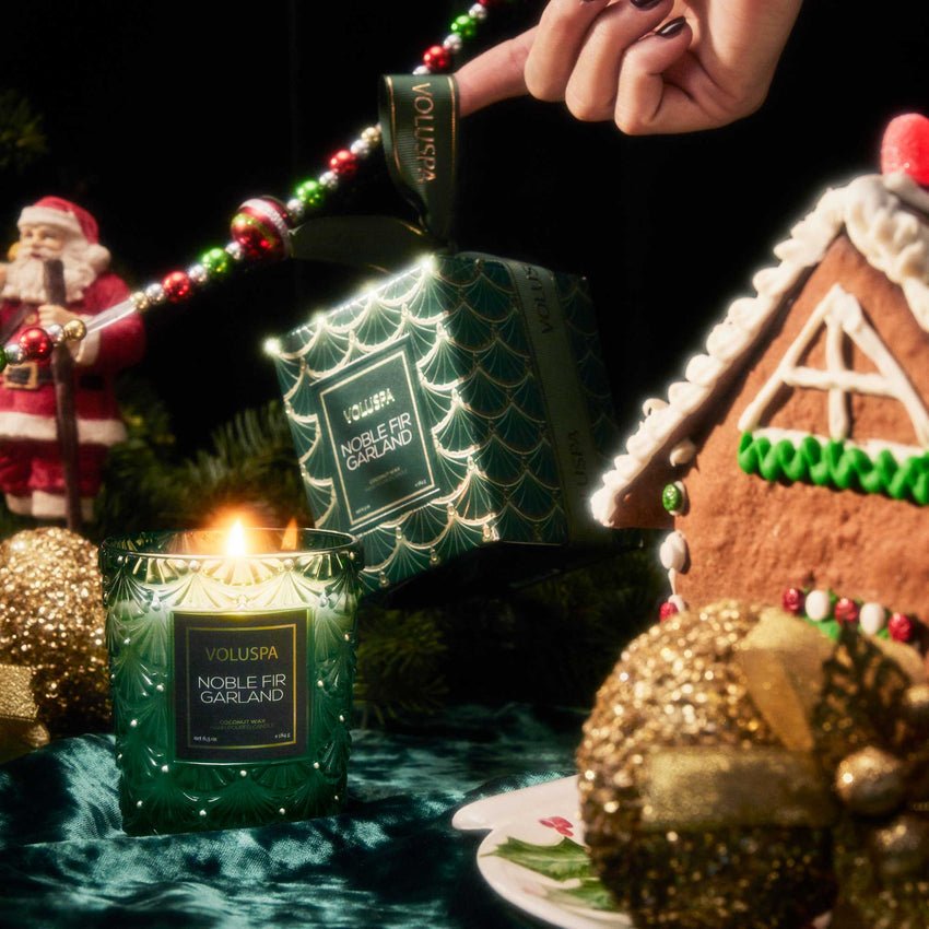 A hand holds a boxed Voluspa - Noble Fir Garland Classic 6.5oz Candle near a glowing candle, gingerbread house, gold ornaments, and Santa figurine, setting a festive scene with the woody aroma of Noble Fir.