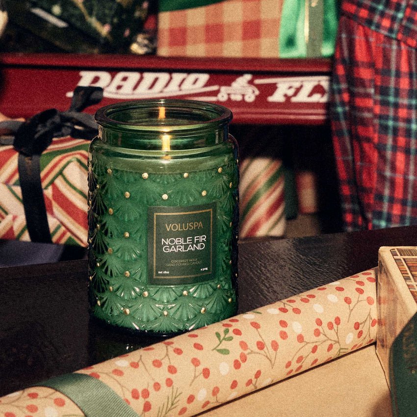 A green Voluspa - Noble Fir Garland Large Jar 18oz Candle emits a woody scent on the table. Wrapped gifts and patterned wrapping paper rolls surround it, with a red Radio Flyer wagon in the background, completing this cozy scene.