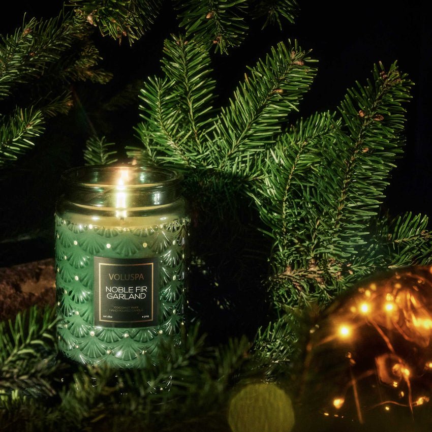 A green Voluspa - Noble Fir Garland Large Jar 18oz Candle is lit, infusing the air with a woody fragrance among evergreen branches. A blurred, glowing gold ornament in the foreground enhances the festive and cozy scene.