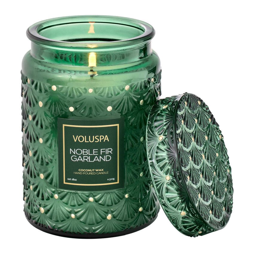 The Voluspa - Noble Fir Garland 18oz candle features a green glass jar and textured fir-branch pattern lid, exuding a woody scent. The coconut wax candles green label with gold text identifies it, as the decorative lid leans gracefully against the jar.