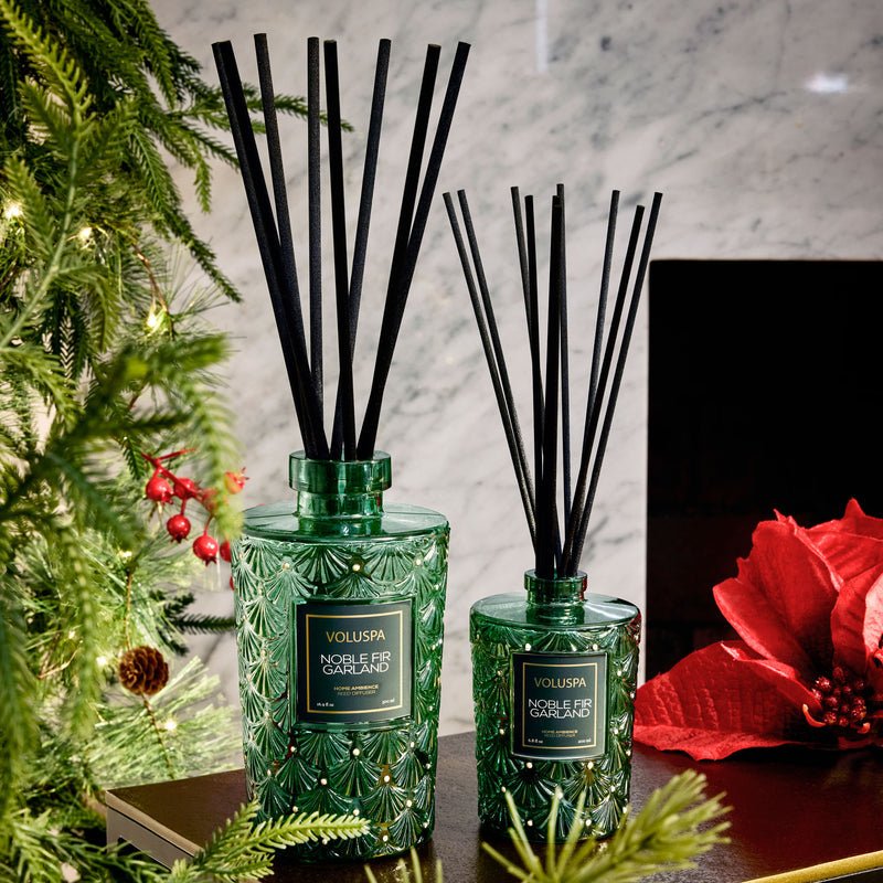 Two Voluspa Noble Fir Garland Reed Diffusers with black reeds sit beside a red poinsettia and ornamented Christmas tree branches. Green glass vessels hold the festive fragrance, set against a marble wall backdrop.