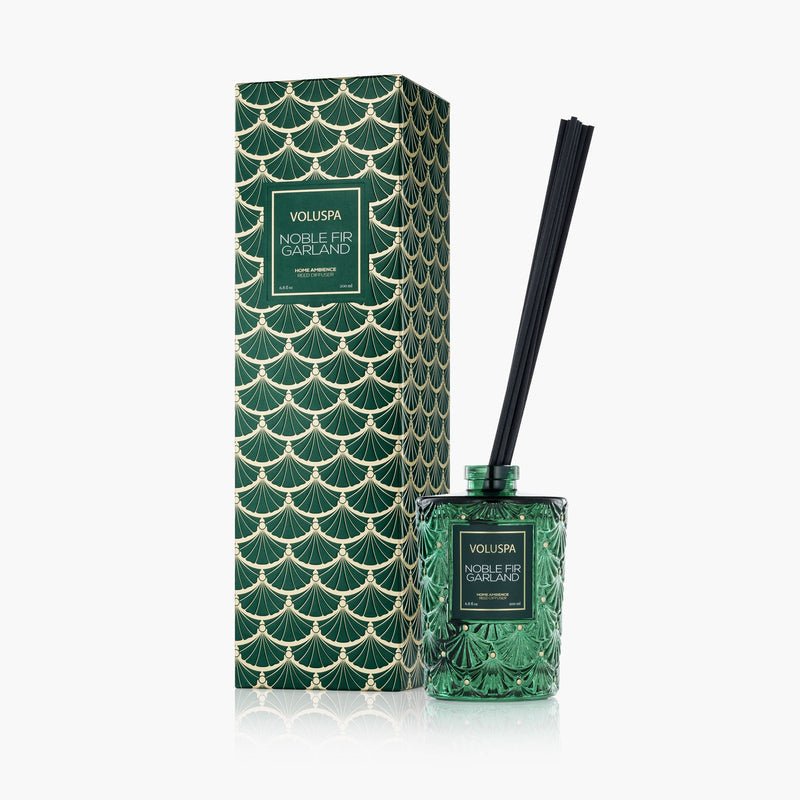 The Voluspa - Noble Fir Garland Reed Diffuser with black reeds and cedarwood oil is shown beside its decorative box, which has a gold and green scalloped design on white.