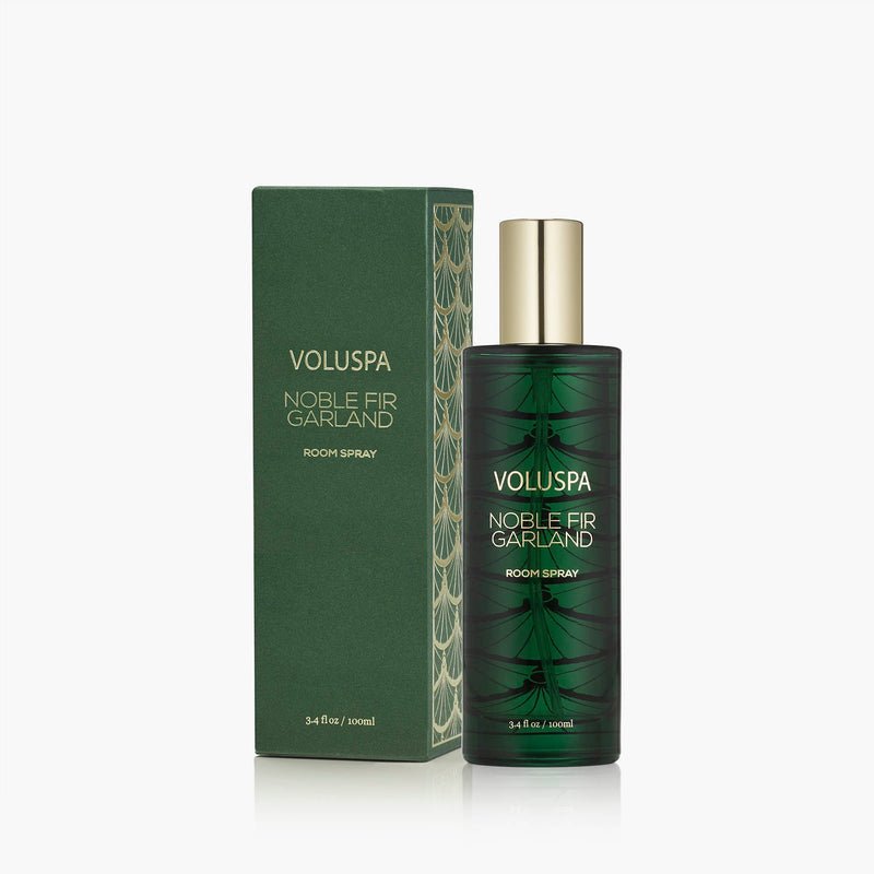 A green Voluspa - Noble Fir Garland Room Spray bottle with gold accents stands beside its matching box, both showcasing elegant, nature-inspired designs—a festive holiday fragrance for any room.