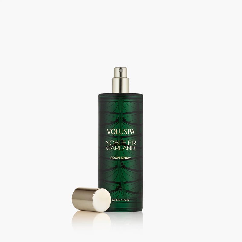 A green Voluspa - Noble Fir Garland Room Spray with a gold cap sits on a white background, decorated with a fir tree design and gold text, evoking the festive scent of the holidays.