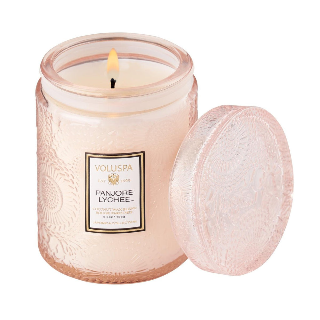 The Voluspa Panjore Lychee 5.5oz candle features a pink-hued textured glass jar with an ornate lid and botanical label. Made from a coconut wax blend, it emits a soft, glowing ambiance when lit.