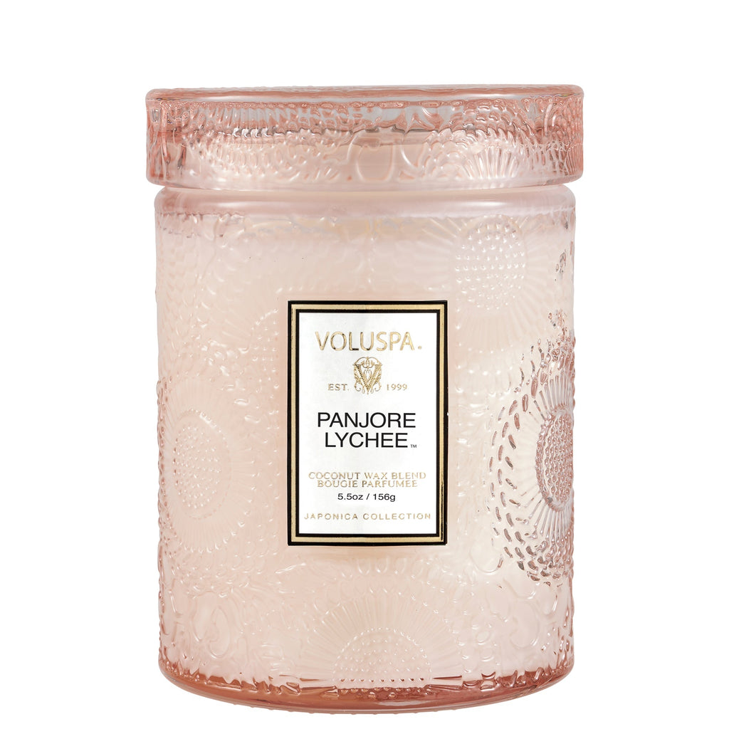 The Voluspa Panjore Lychee Small Jar Candle from the Japonica Collection is a vegan, textured pink glass candle with a floral pattern and coconut wax blend. It has fragrant notes of lychee and features a label stating Voluspa, Est. 1999, Bougie Parfumée, 5.5oz/156g, combining elegance with sustainability.