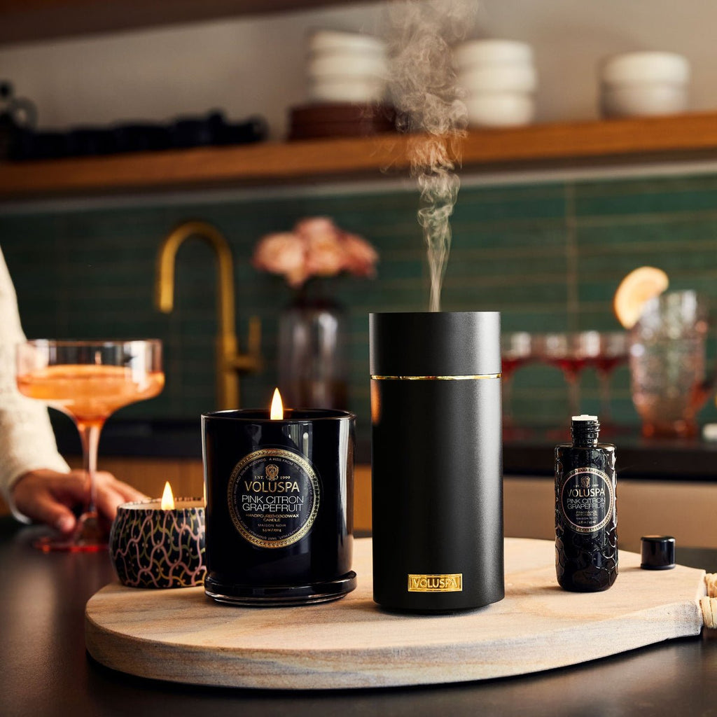 A kitchen counter features a lit black candle and a sleek black vaporizer subtly releasing Voluspa - Pink Citron Grapefruit fragrance. A chic black bottle with gold labels adds elegance, while a blurred figure and a champagne glass create a stylish backdrop.