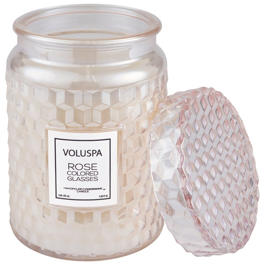 A Voluspa Rose Colored Glasses large 18oz jar candle, made from a coconut wax blend, features a white candle in a geometric glass jar. The lid is off, and it emits a delightful floral fragrance.