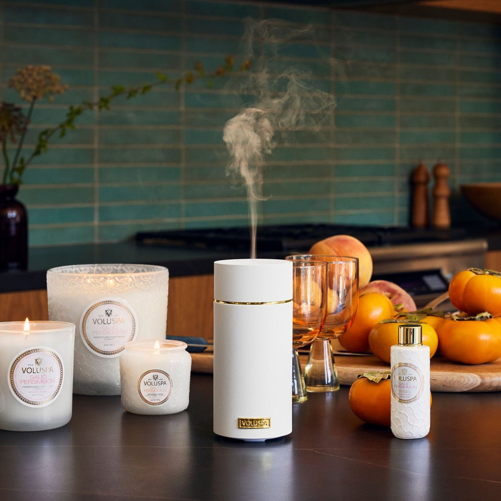 In a cozy kitchen, a Voluspa diffuser emits Saijo Persimmon fragrance oil mist. Its surrounded by Voluspa candles and a fragrance bottle, with persimmons and wine glasses in the background against teal tiles and the stove.