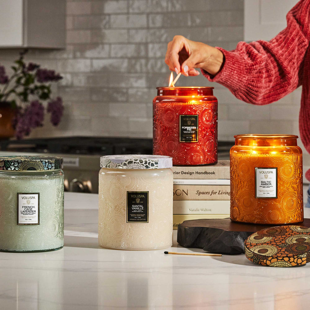 A hand lights a Voluspa - Santal Vanille Luxe Jar 44oz Candle on a kitchen island, filling the room with its warm scent. Nearby, three other candles—one green and two ivory—rest among a book and decorative items, enhancing the kitchens charm with its tiled backsplash and stainless steel accents.