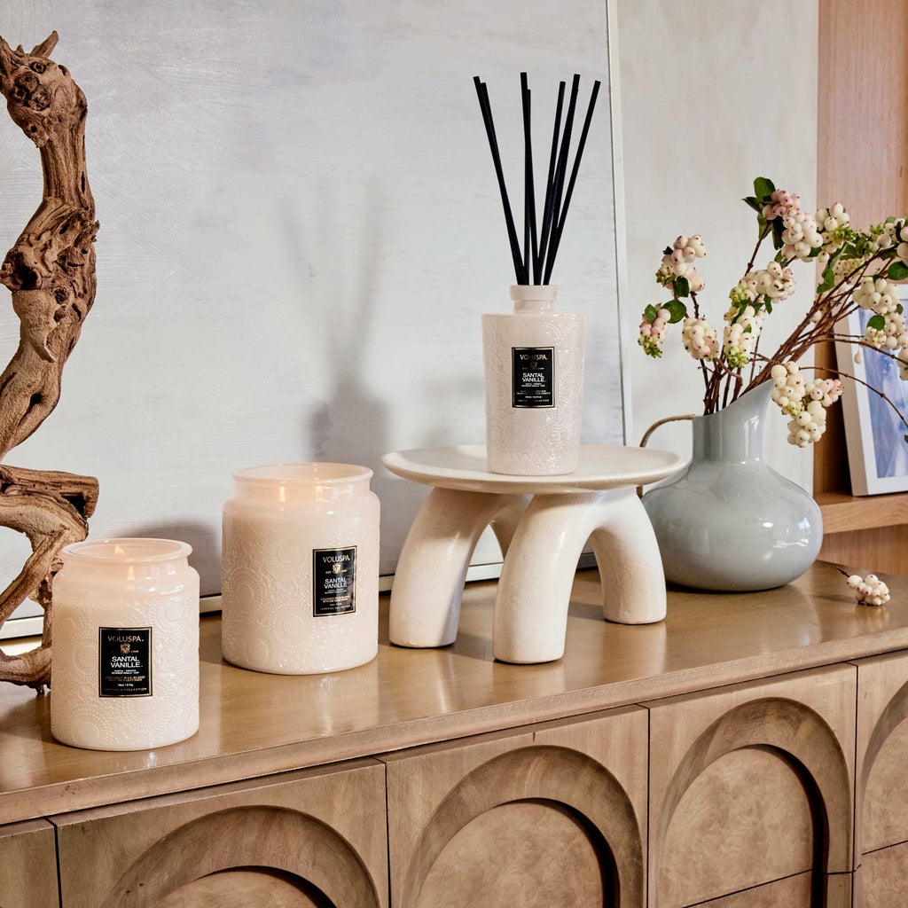 On a wooden console, the Voluspa Santal Vanille Luxe Jar candle is lit alongside a reed diffuser on a curved stand, driftwood, and a vase with white flowers against a soft, neutral-toned wall.