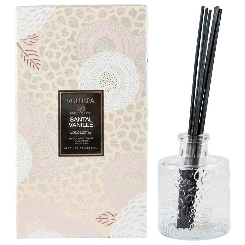 A decorative glass diffuser with black reeds and a Voluspa Santal Vanille label sits beside a floral-patterned Japonica Collection box, blending sandalwood and vanilla.