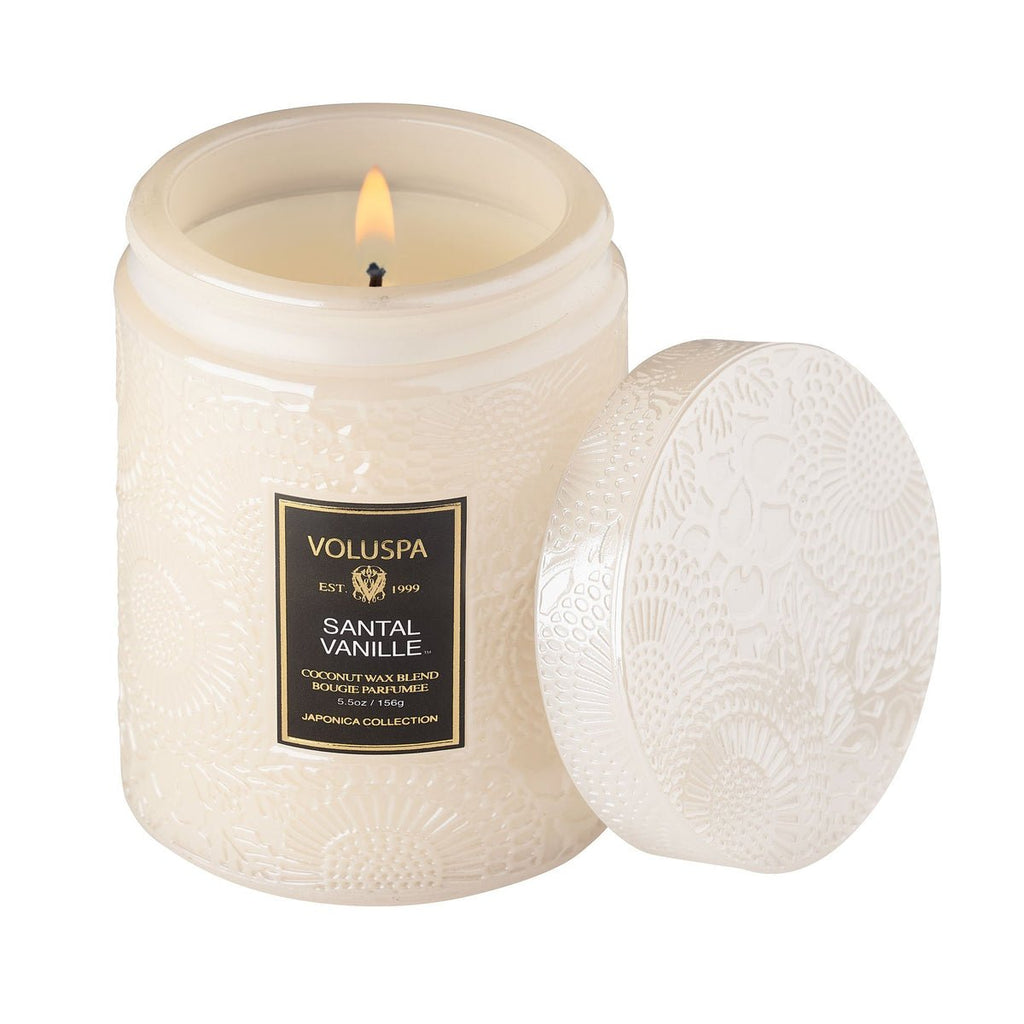 The Voluspa - Santal Vanille Small Jar 5.5oz Candle fills the room with sandalwood and vanilla notes. Its lit white candle sits in an embossed glass jar, with the matching lid elegantly resting beside it.