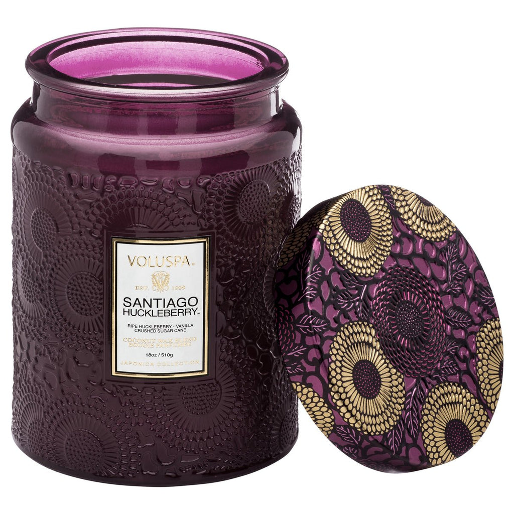 The Voluspa - Santiago Huckleberry Large Jar 18oz Candle features a decorative purple glass jar with an embossed floral pattern and is made from coconut wax for a cleaner burn. The colorful floral design on the lid leans against the jar.