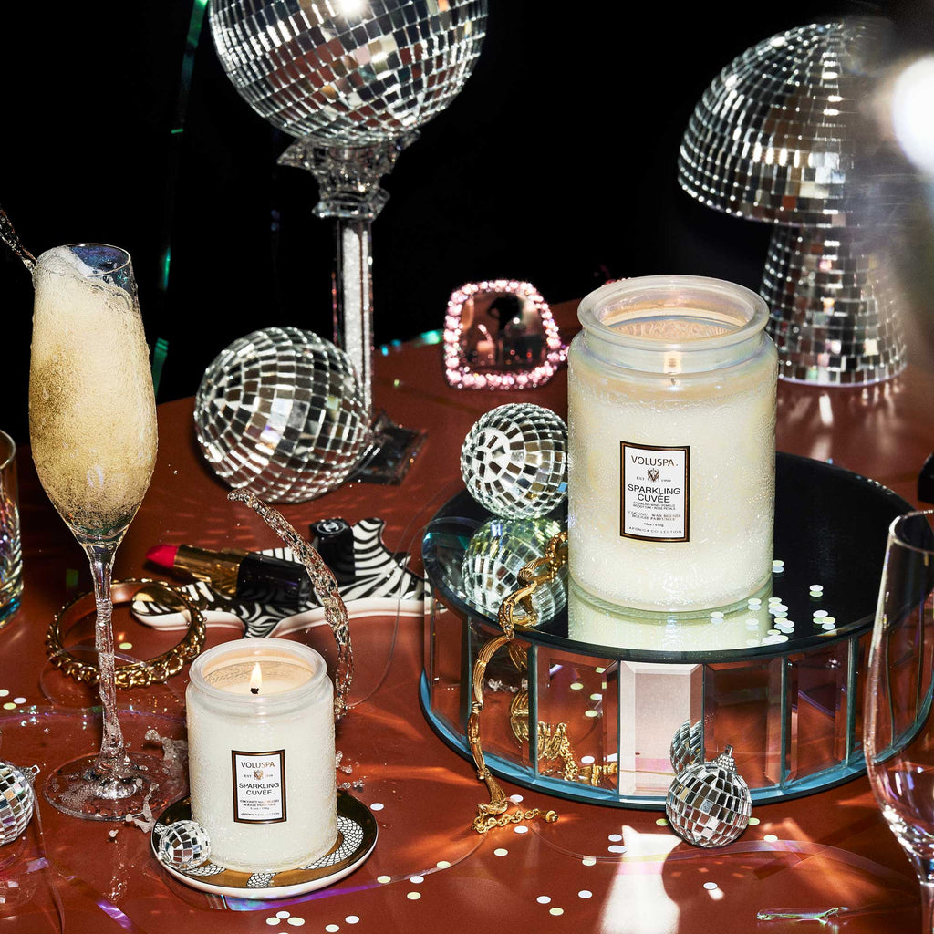 A celebratory table features a Voluspa Sparkling Cuvée candle in an 18oz jar, a champagne flute, disco balls, confetti, makeup items, and the glow from two coconut wax blend candles reflecting on the glossy surface.