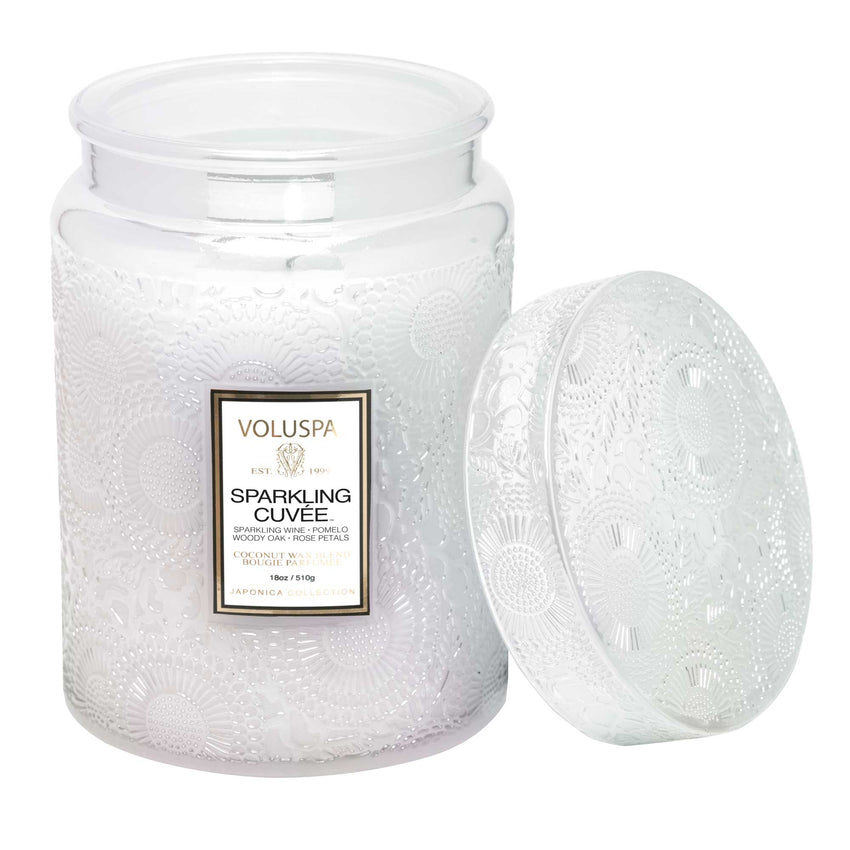 An 18oz Voluspa - Sparkling Cuvée candle, crafted from a coconut wax blend, sits in a decorative glass jar with intricate patterns and is complemented by a matching lid. The gold-accented label and brand logo enhance its luxurious fragrance appeal.