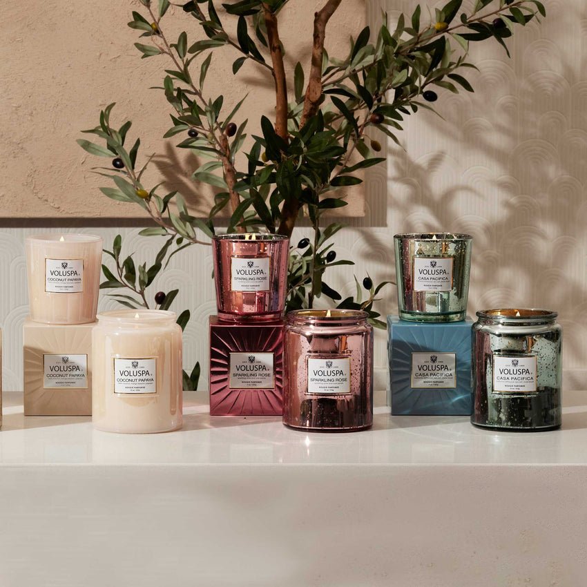 Nine Voluspa candles, including the Sparkling Rose Large Jar 18oz Candle, in pastel and metallic hues rest on a white surface. Each coconut wax candle is in a glass jar with a label. Greenery accents the backdrop, enhancing the floral fragrance ambiance.