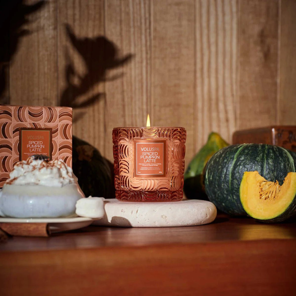 Voluspa’s Spiced Pumpkin Latte Classic 12oz Candle sits on a wooden table with its decorative box, alongside a green pumpkin, pumpkin slice, and whipped cream-topped mug. Its coconut wax blend fills the space with cozy autumn vibes.
