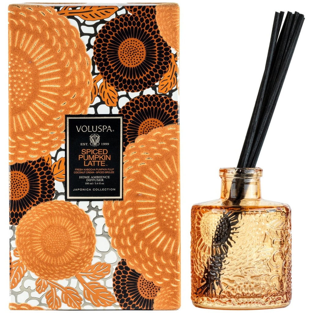 An ornate amber glass diffuser, filled with Voluspa Spiced Pumpkin Latte oil and dark reeds, stands next to a box featuring an intricate orange and black floral design, encapsulating the cozy fall essence.