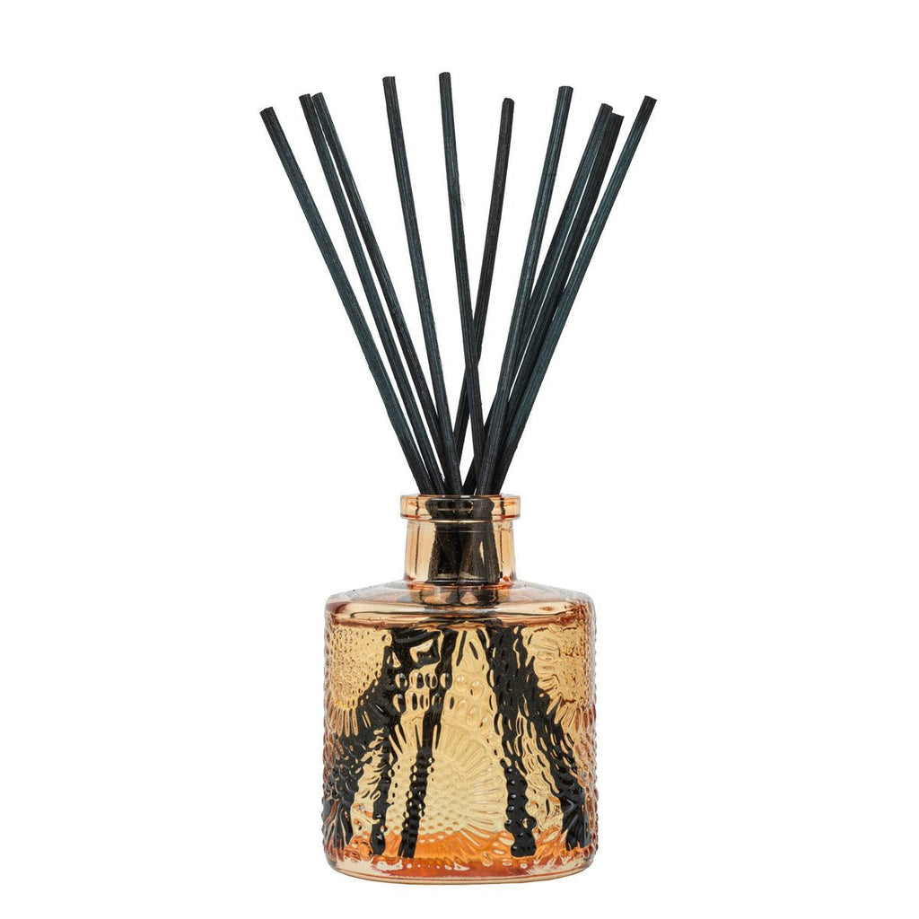 The Voluspa Spiced Pumpkin Latte Reed Diffuser features a textured amber glass bottle filled with fragrant oil, releasing a cozy scent of spiced pumpkin latte through black reeds that fan out to warm your home.