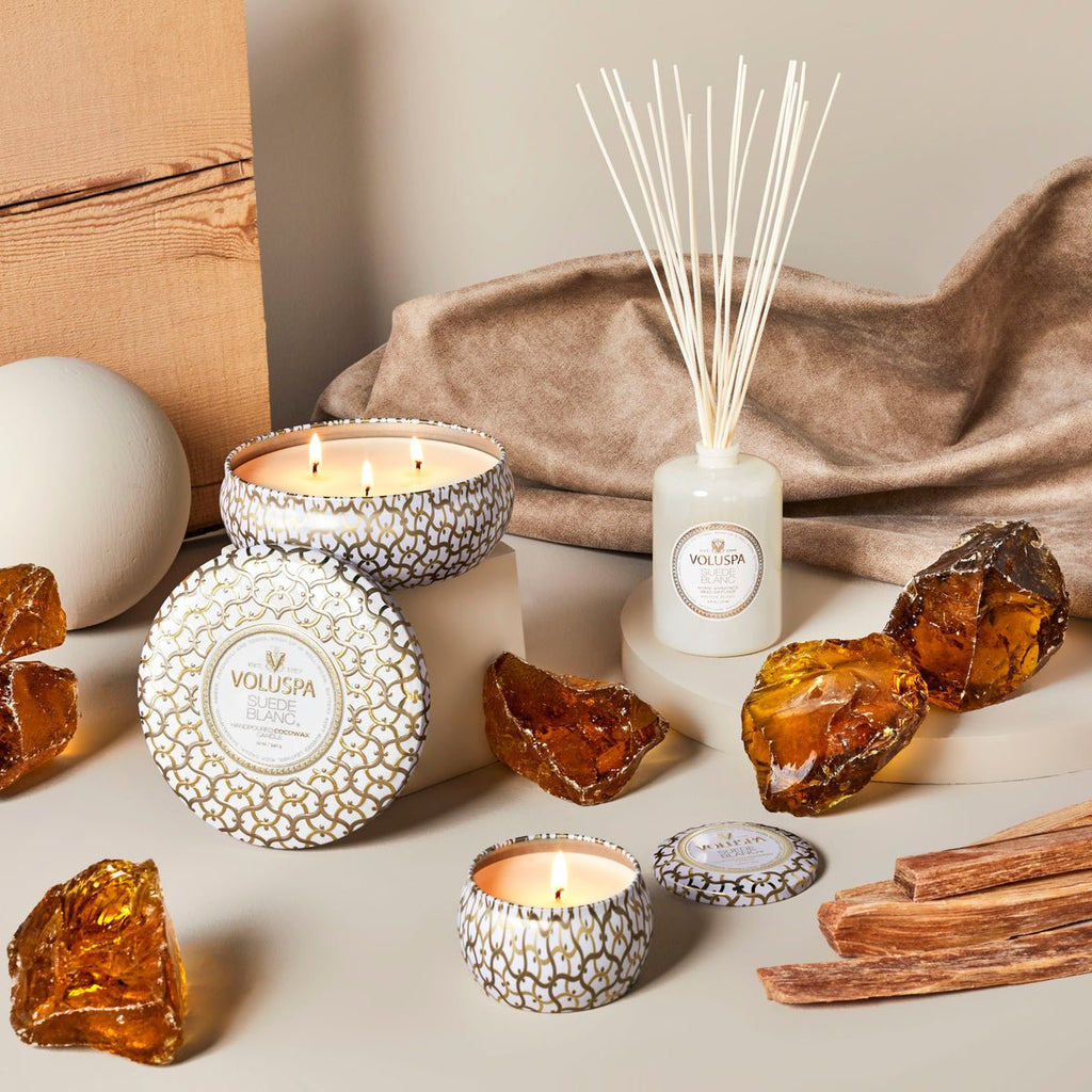 Voluspas Suede Blanc 172ml Reed Diffuser is artfully arranged with candles in decorative tins, amber-like stones, and wooden sticks on a neutral surface. A soft fabric backdrop adds warmth and texture, creating an inviting fragrance scene without the flame.