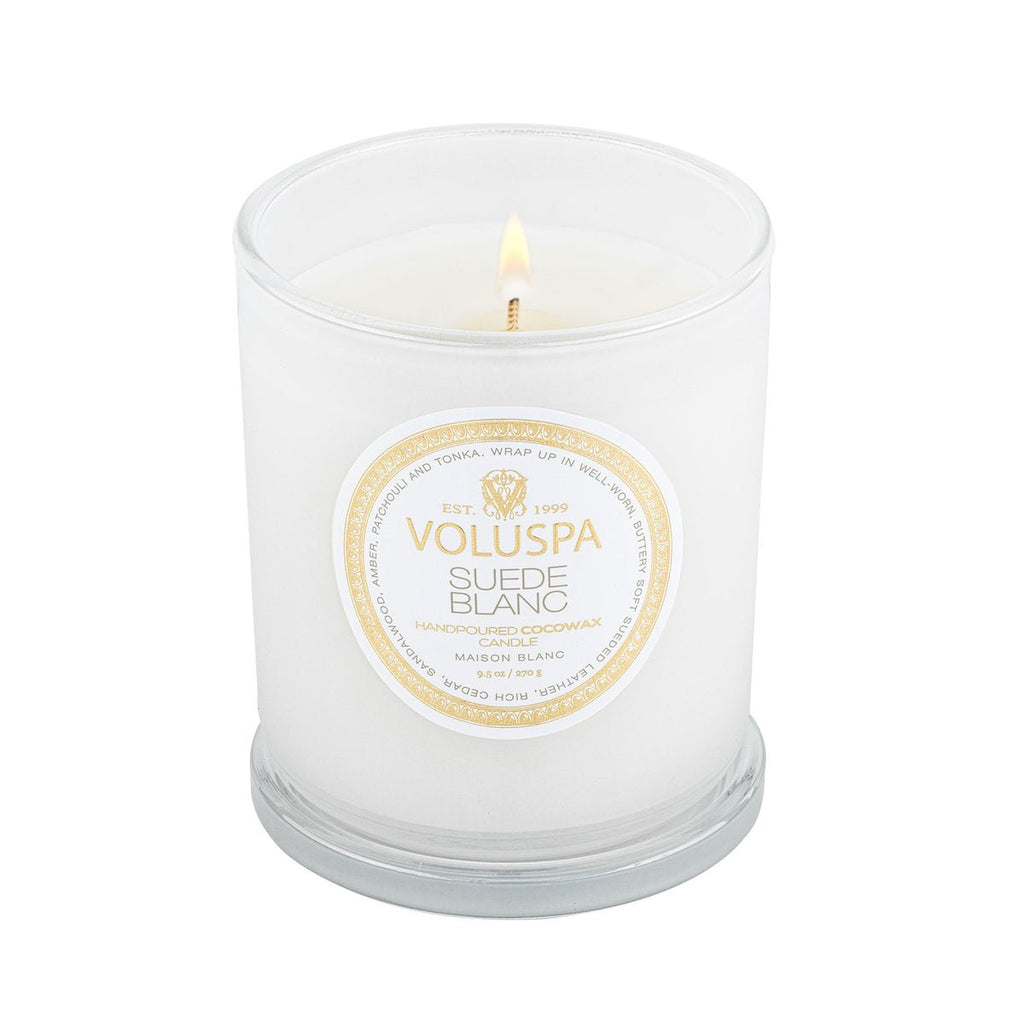 A lit Voluspa Suede Blanc Classic 9.5oz Candle, in a clear glass with white wax, features gold and black text on its label. Its a handcrafted, phthalate-free coconut wax blend with buttery sueded leather hints. The flame softly illuminates the simple setting.