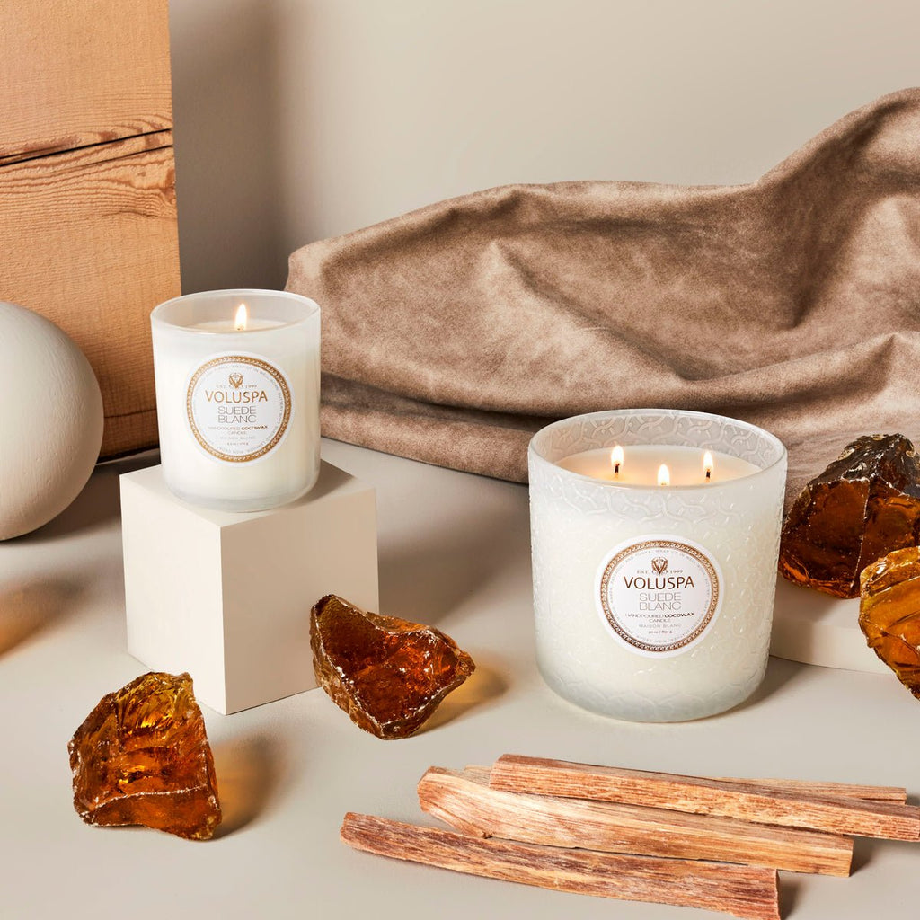 Two Voluspa Suede Blanc Classic candles in phthalate-free white jars complement amber resin and wood sticks on the table, while a textured beige cloth and a wooden board enhance the coconut wax scent for a cozy atmosphere.