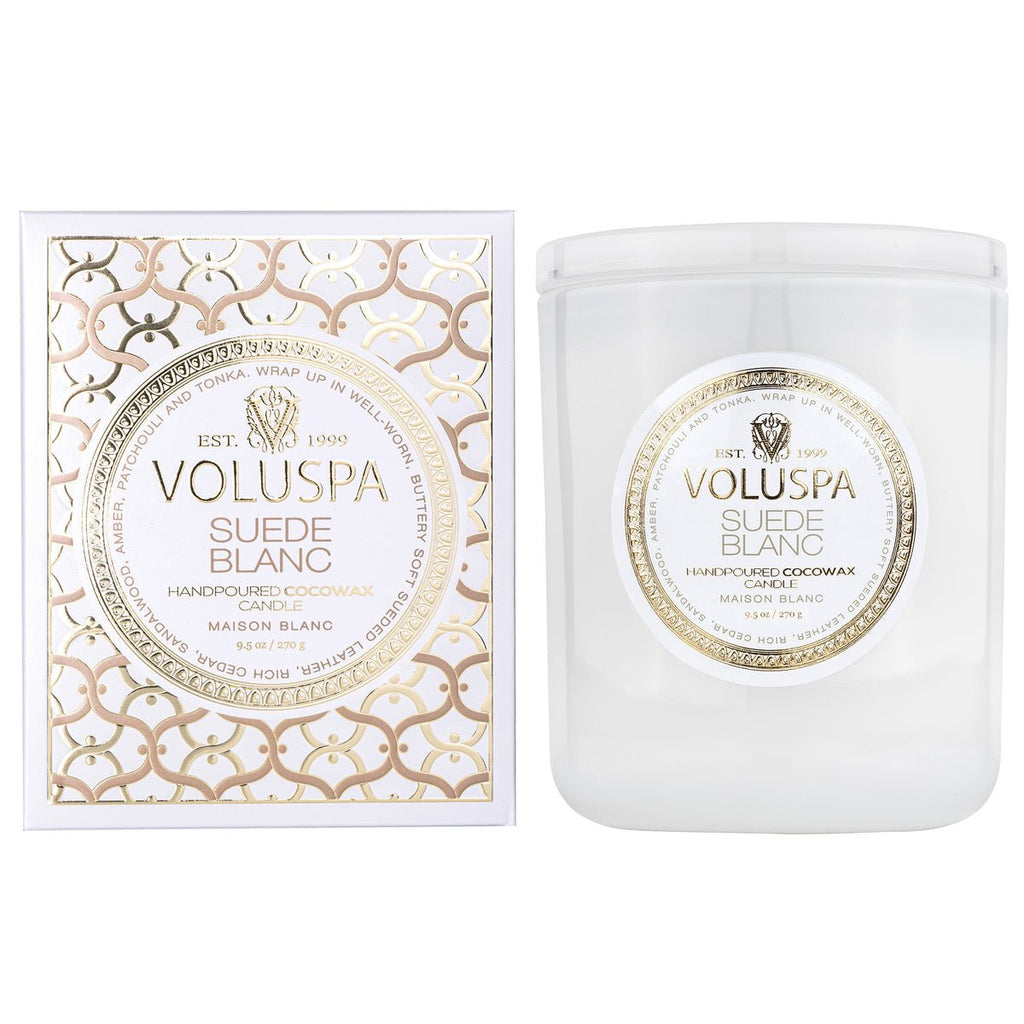 Voluspas Suede Blanc Classic 9.5oz candle, made from a coconut wax blend, sits in a clear glass jar next to its decorative box with a gold and white geometric design, evoking fresh sueded leather.