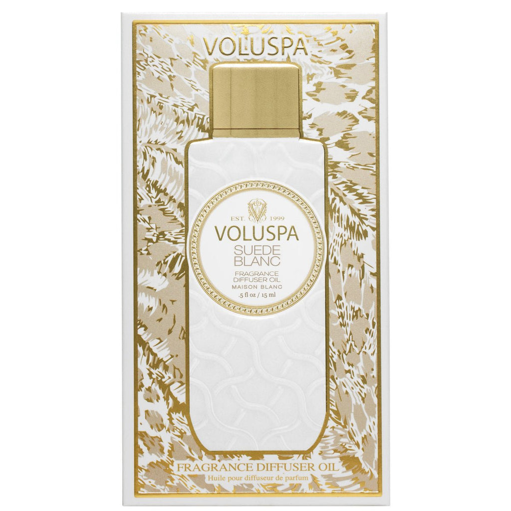 The image shows a Voluspa - Suede Blanc Ultrasonic Diffuser Oil box adorned with a gold and white floral pattern. The round Voluspa logo stands at the center, surrounded by elegant text describing the exquisite sueded leather-inspired fragrance.