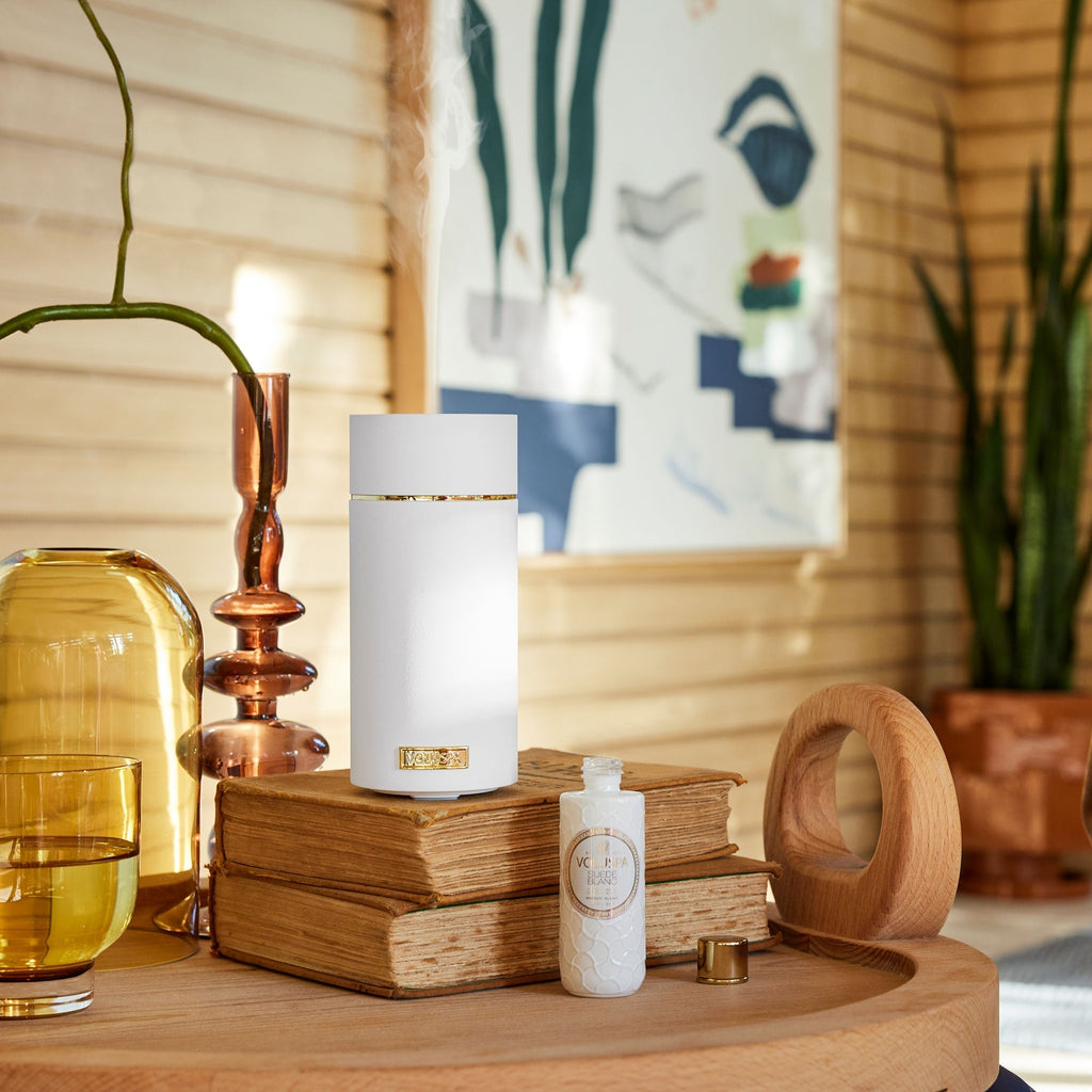 A white Voluspa diffuser with Suede Blanc oil emits mist over stacked books on a round wooden table, alongside a small bottle, yellow glass jar, and brown candlestick. A wooden wall features abstract art in the backdrop, creating an inviting scene.