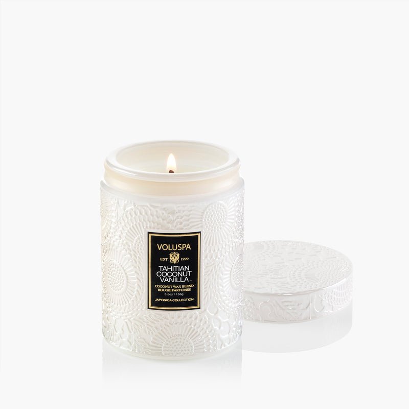 A white embossed glass Voluspa - Tahitian Coconut Vanilla 5.5oz Small Jar Candle, featuring a black and gold Voluspa label, is lit beside its matching decorative lid on a white background.