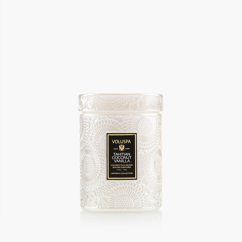 A white glass Voluspa jar candle (5.5oz) features embossed floral patterns, a clear lid, and a gold-and-black label reading "Voluspa - Tahitian Coconut Vanilla." The background is plain white.