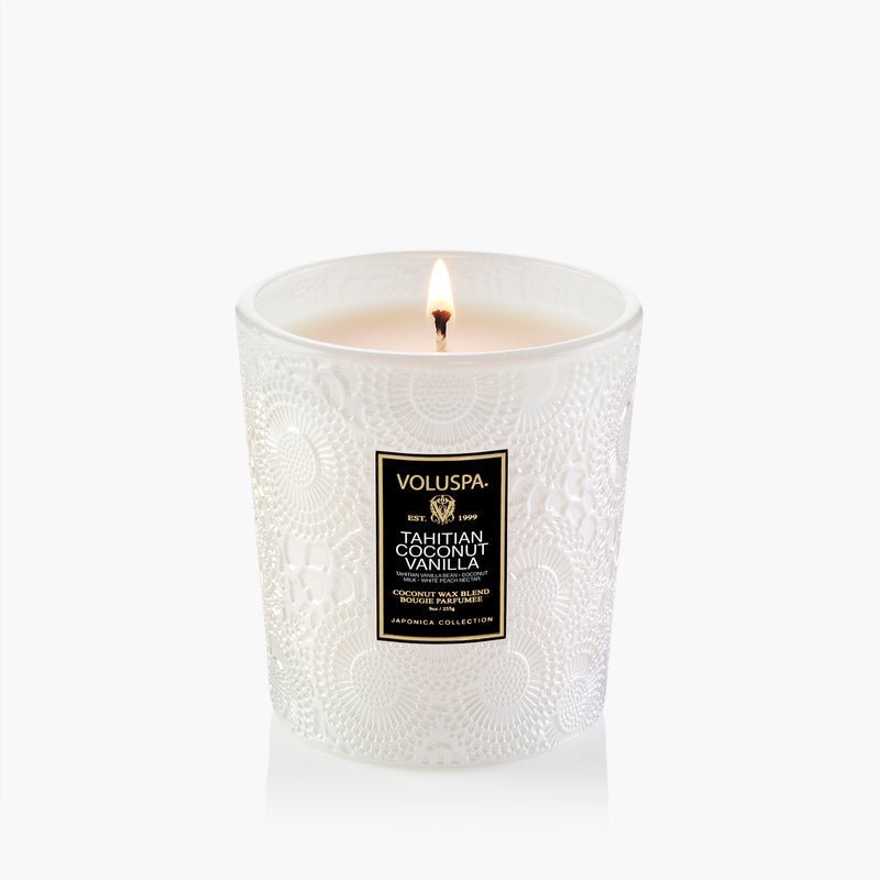 A lit Voluspa - Tahitian Coconut Vanilla Classic 9oz Candle with an embossed glass holder and black-and-gold label sits against a plain white background.