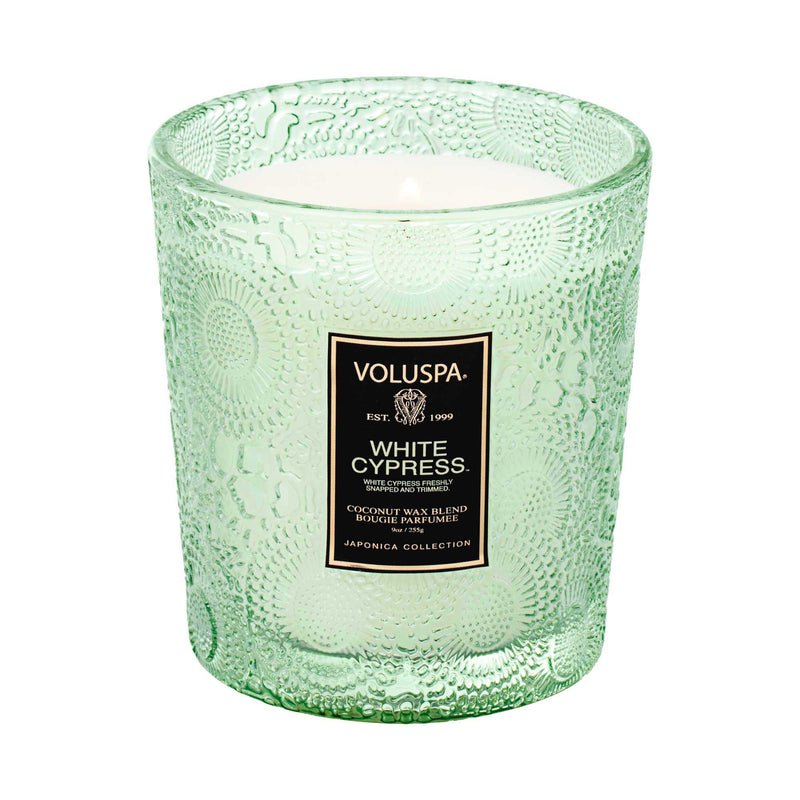 The lit Voluspa - White Cypress Classic Candle is a green textured glass candle from the Japonica Collection, featuring a clean-burning coconut wax blend.