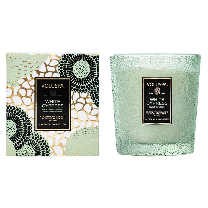 The Voluspa - White Cypress Classic Candle 9oz features an elegant textured green glass container and a clean-burning coconut wax blend. It comes with a decorative box in intricate black and gold patterns on a light green background, perfect for adding elegance to any space.