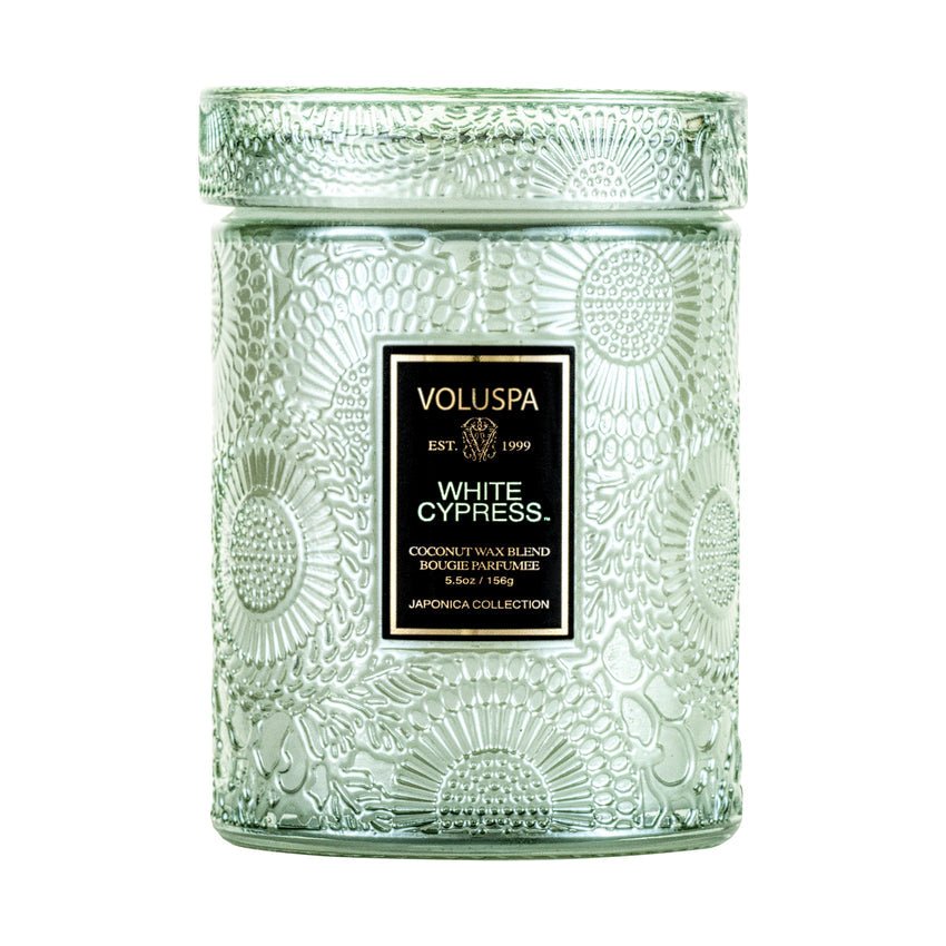 The Voluspa - White Cypress Mini Jar 5.5oz Candle is a holiday scent in a decorative glass jar with an embossed floral pattern, featuring a luxurious coconut wax blend and a black label accented by gold text and trim.