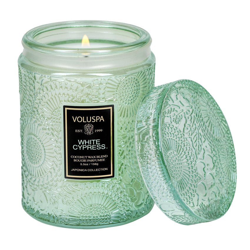 The Voluspa - White Cypress Mini Jar Candle flickers in a green embossed glass jar with its lid open beside it. The label, reading Voluspa White Cypress, features intricate patterns. Part of the Japonica Collection, this holiday scent has a luxurious coconut wax blend.