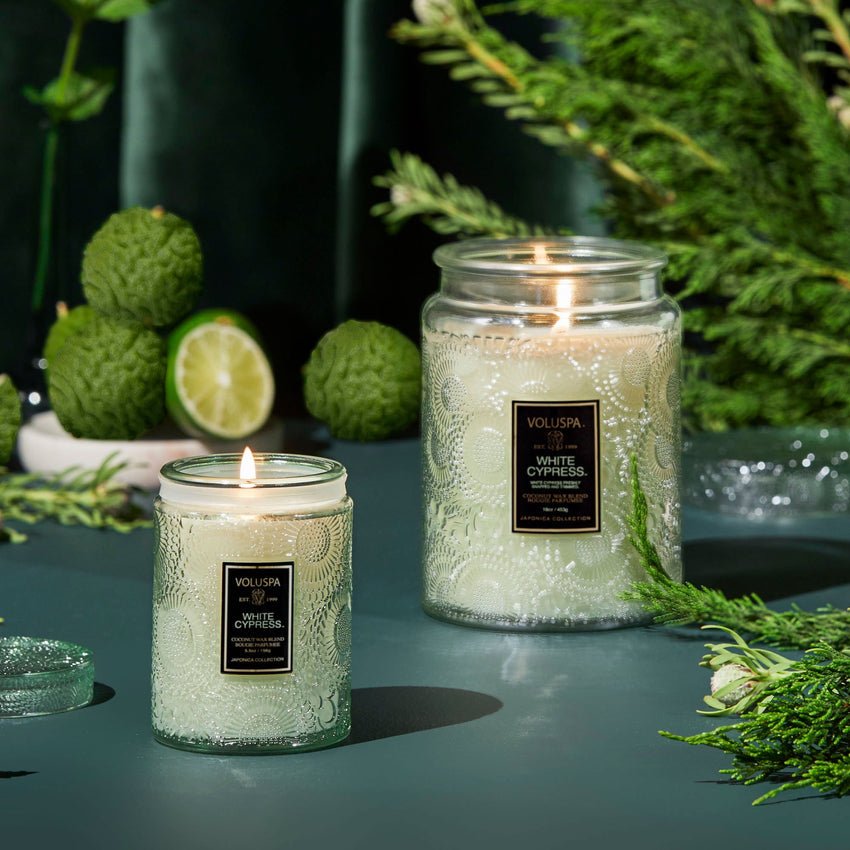 Two Voluspa White Cypress mini jar candles, crafted from luxurious coconut wax, sit on a green surface. Encircled by sprigs of greenery, a lime slice, and textured green spheres, these holiday-scented candles feature Voluspa labels.