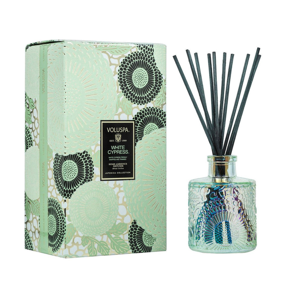 A decorative, fragrant Voluspa - White Cypress Reed Diffuser with black reeds stands beside a patterned box. The bottle and box share intricate floral designs in a soothing teal color, evoking the fresh scent of evergreen wood.